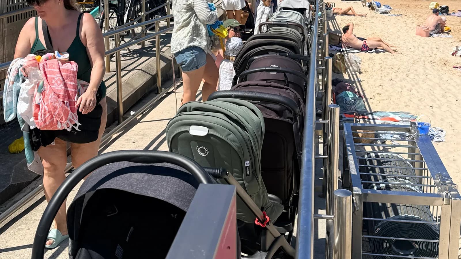 A long line of prams parked nose-to-tail along the handrail of the ramp at the northern end of Bondi Beach, with the sand and the ocean pool behind