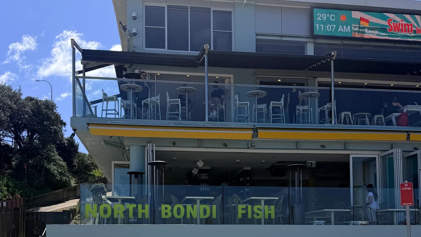 The North Bondi surf club building and the North Bondi Fish terrace above the promenade, with people at the upstairs tables on a clear summer morning