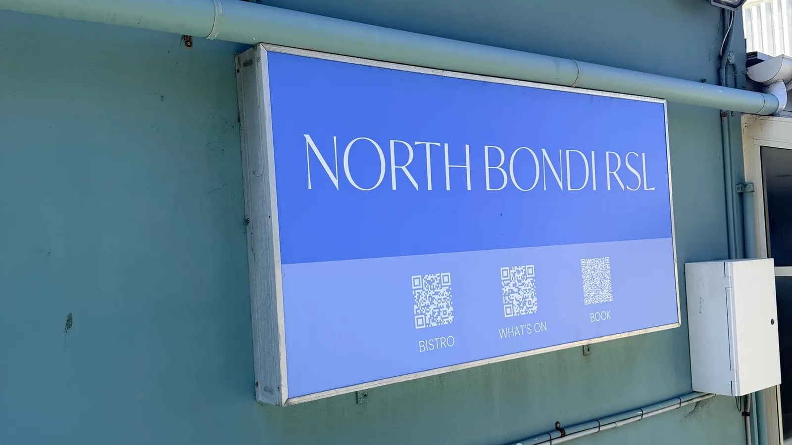 The North Bondi RSL sign on the pale green exterior wall of the club, with QR codes below it labelled bistro, what's on and book